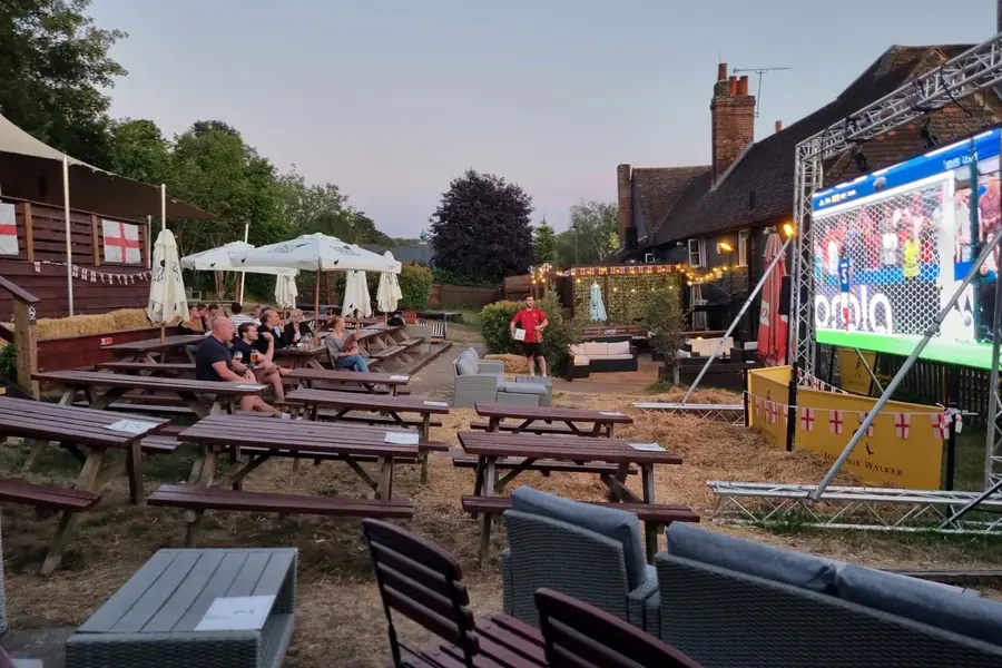 The garden filling up on a summer evening, picnic benches in front of a big screen showing football