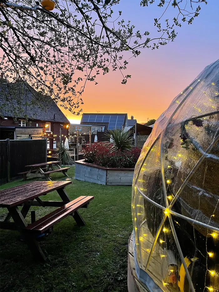 A pod in the garden at sunset, lights on, a picnic bench beside it